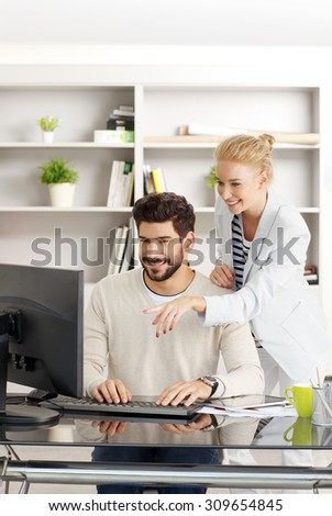 Portrait of two young business people working together at office. Professional man sitting in front of monitor and typing presentation while creative female standing next to him and consulting.