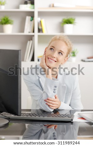 Portrait of thinking businesswoman sitting at desk in front of computer. Young assistant wearing casual clothing and smiling.