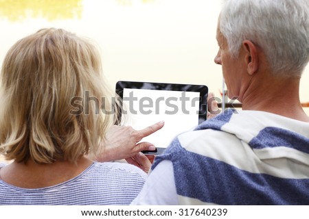 Portrait of senior couple sitting at lakeside. Elderly woman holding hand white blank screen digital tablet while old man sitting next to her and looking the display.