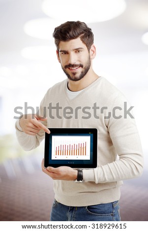 Portrait of young sales man standing at workplace and holding hand digital tablet with column chart display. Businessman point out the tablet display.
