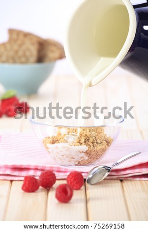 Pouring milk on a bowl of cereal (muesli/granola) with fresh strawberries, raspberries and fruit in the background.