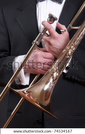 A slide trombone is held in front of a tuxedo clad male torso.