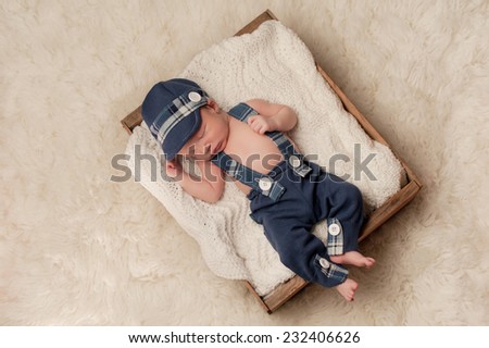 Overhead shot of an eight day old newborn baby boy wearing a blue newsboy cap, suspenders and pants. He is sleeping in an old wooden crate. Shot in the studio on a cream colored flokati rug.