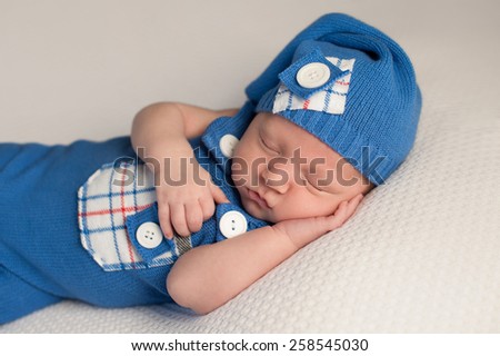 A sleeping newborn baby boy. He is wearing an upcycled blue and white romper and matching cap.