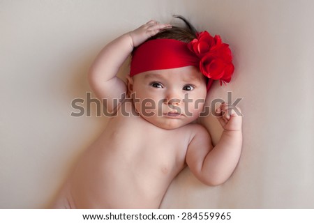 A portrait of an alert, 3 month old baby girl wearing a large, red, flower headband. She is lying on her back on a cream colored blanket and has a funny expression on her face.