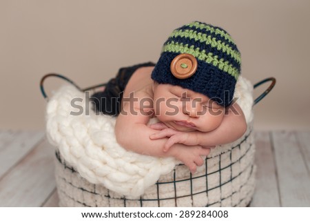 Three week old newborn baby boy wearing jeans and a crocheted blue and green beanie hat. He is sleeping on his stomach in a wire basket.