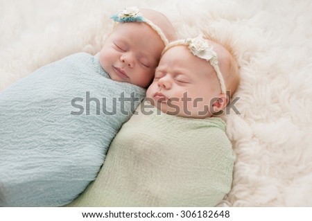 Seven week old fraternal, twin baby girls swaddled and sleeping on a white flokati rug. One sister is smiling.