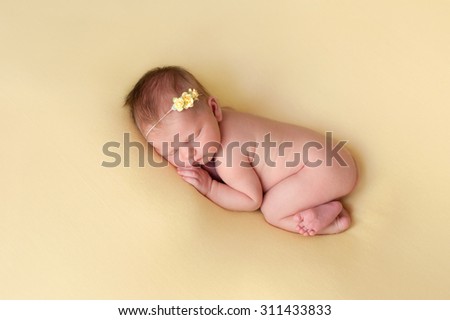 A portrait of a beautiful nine day old baby girl sleeping in a curled up fetal position on a yellow blanket. She is wearing a yellow rose headband.