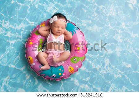 Nine day old newborn baby girl sleeping on a tiny inflatable swim ring. She is wearing a crocheted pink and white bikini.