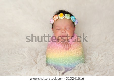A sleeping nine day old newborn baby girl bundled up in a rainbow colored swaddle. She is propped up on a cream colored flokati (sheepskin) rug and wearing a crown made of roses.