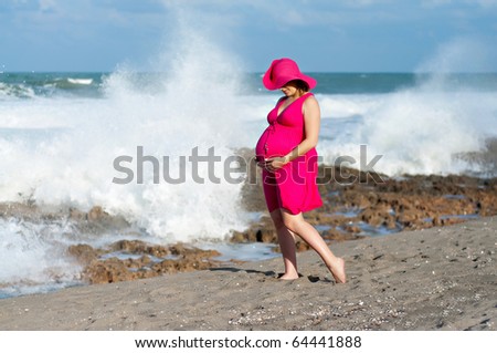 A beautiful pregnant woman standing on the beach in a pink sundress and hat with the ocean waves crashing behind her.