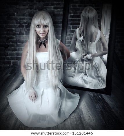 Young beautiful blond woman sitting on wooden floor in old dark room with big knife in mirror reflection