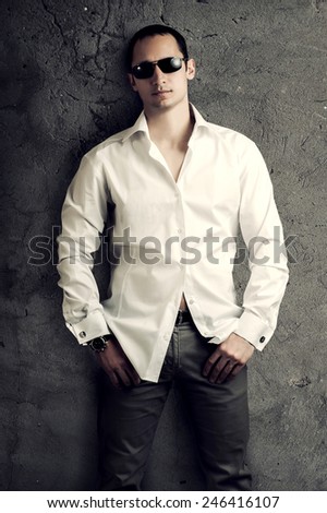 Fashion portrait of young sexy man wearing white luxury shirt and sunglasses poses about gray wall