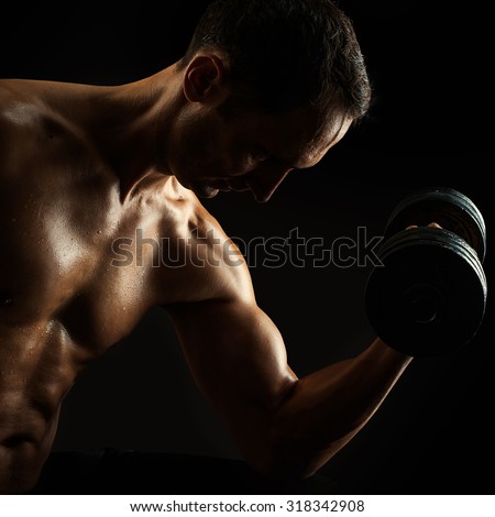 Dark contrast brutal silhouette of young muscular fitness man. Bodybuilder with beads of sweat training in gym. Working out with dumbbells on black background