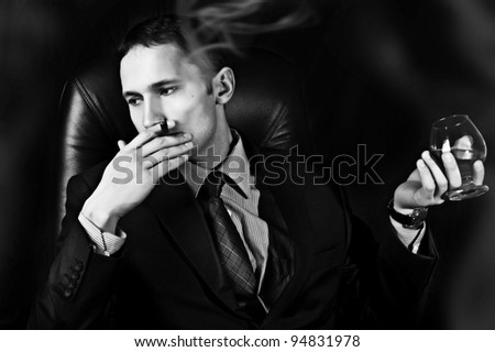 Black and white portrait of young handsome business man with Old Brandy Glass in hand and smoking cigar on black background. men\'s club