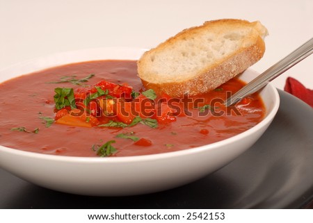 A side view of a bowl of tomato, red pepper and basil soup with bread