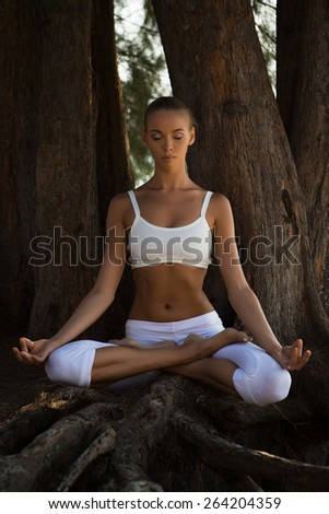 Pretty slim Woman in white costume doing Yoga pose near big tree