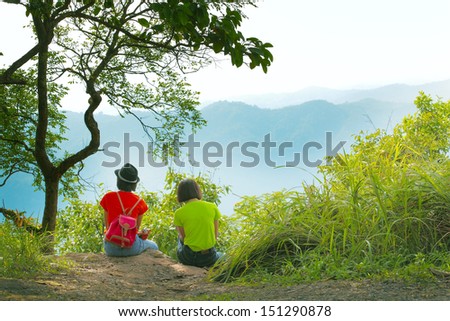 Women in red and green t-shirt sitting at the cliff looking at mountain scenery far away, Thailand