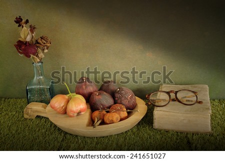 Still life art photography on fruits set with old book glasses and dry roses in vase over grunge backdrop and artificial grass floor
