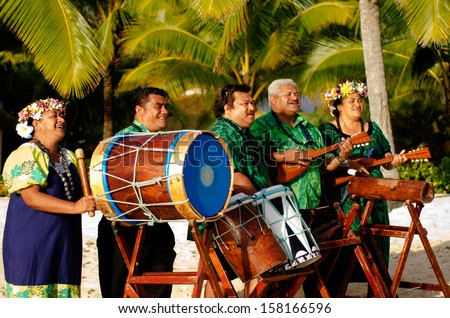 Group portrait of Polynesian Pacific Islanders band plays Tahitian music on tropical beach with palm trees in the background.
