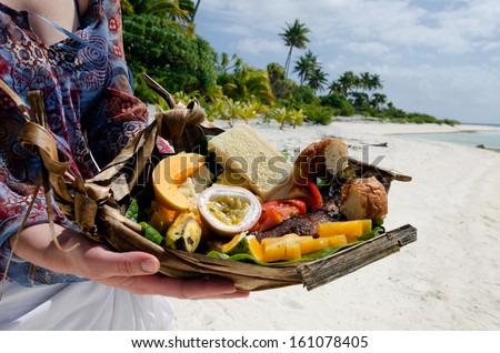 Young woman hands carry tropical food of grilled fish, fruits and vegetables dish served on deserted tropical island in Aitutaki lagoon, Cook Islands.