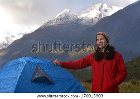 Portrait of a young woman camping outdoors under a high snow-capped mountain during sunset.