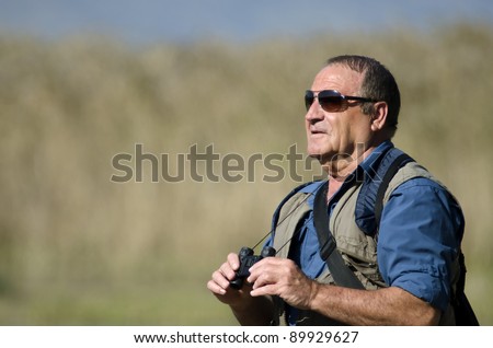 A senior hiker and bird watcher is searching for birds with binoculars.