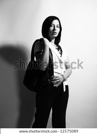 A black and white high-key portrait about a pretty trendy lady with black hair who is looking ahead and she has an attractive look. She is wearing black pants, a white coat and a stylish handbag.