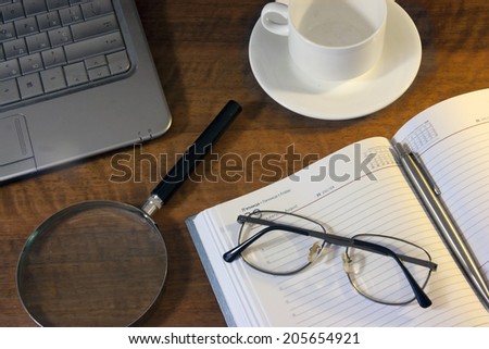 Wooden desk with an appointments diary, pen, glasses, magnifying glass and a coffee cup on it