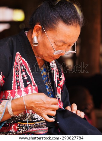 Chiang Mai, Thailand - March 07, 2011 - Portrait of 60 year old Thai woman, embroider a piece of cloth in front of her bamboo house