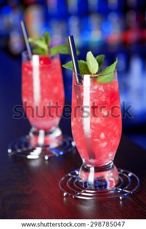 Two glasses of Eclipse cocktail at the bar, lit in blue light. Ingredients: 3 oz gin 1 oz raspeberry 1/2 grenadine crushed ice to the top mint and maraschino cherry for decoration