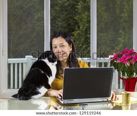 Mature Asian woman and family cat cuddling while working at home with large windows in background