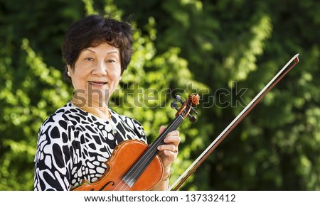 Horizontal photo of Senior Asian woman taking a break from playing the violin outdoors with bright green trees in background