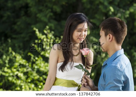 Horizontal photo of a young adult man holding pink rose with his lady friend looking into his eyes with green trees in background