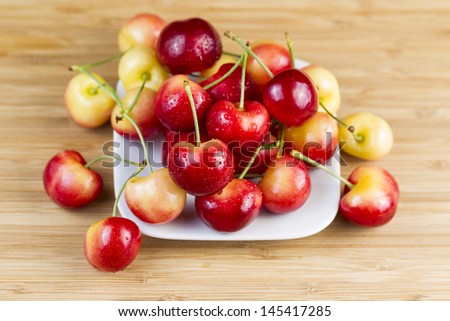 Horizontal photo of Rainier cherries, with water drops, in white dish on natural bamboo board