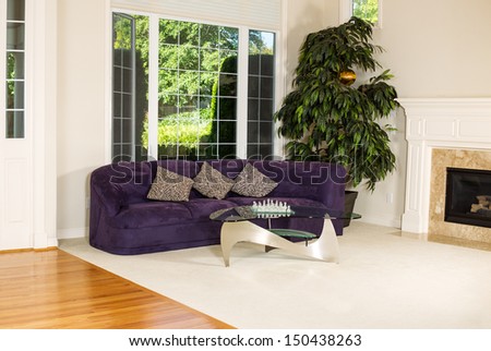 Horizontal photo of formal living room with suede leather couch, glass table, carpet, large windows, plant, fireplace and hard wood floors