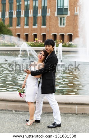 Vertical photo of young adult couple relaxing and holding each other with water fountain, flowers, trees and brick building in background