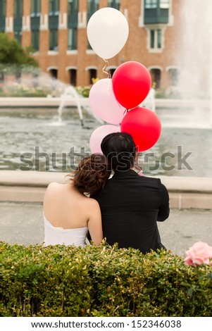 Vertical photo of young adult couple holding balloons while looking at water fountain with trees, flowers in background