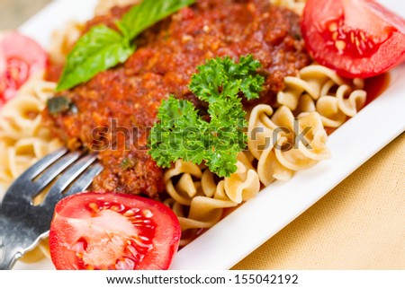 Closeup horizontal photo of freshly cooked pasta, fork, tomato sauce, tomatoes, parsley, basil in a white plate with textured cloth napkin on side of dish