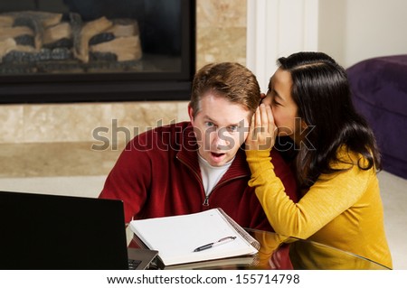 Photo of mature couple, with woman whispering into ear of man who is shocked, while working from home with fireplace and partial sofa in background