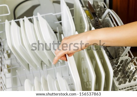 Horizontal photo of female hand putting in dinner dishes in lower dish rack that is filled with white plates, stainless steel knifes, and spoons with red oak cabinet in background
