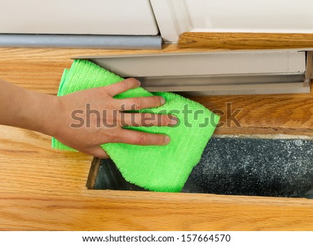 Horizontal photo of female hand cleaning, using microfiber rag, underneath grill plate of heater floor vent with Red Oak Floors and front door in background