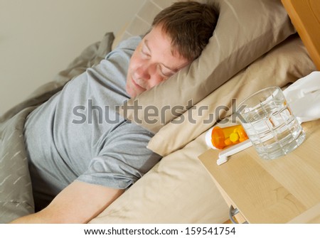 Closeup photo of medicine, thermometer, glass of water and tissues on night stand with mature man bedridden in background