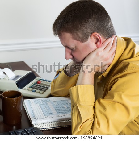 Photo of mature man, holding head with hands, working on his taxes with tax booklet and office equipment in background