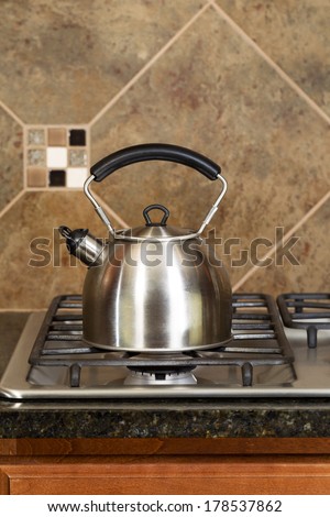 Vertical photo of a stainless steel tea pot on stove top with stone counter tops and tile back splash