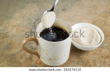 Horizontal photo of a spoon of sugar being poured into a cup of black coffee with stone counter top underneath