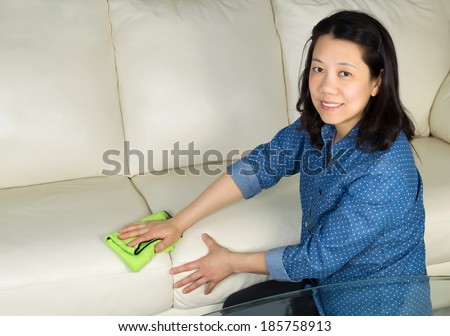 Horizontal photo of mature woman, looking forward, cleaning white leather sofa with microfiber rag in hand