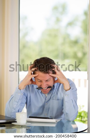 Vertical image of mature man showing extreme stress by biting his pen while working from home with bright daylight coming in from window in background
