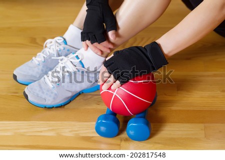 Horizontal image of female hands wearing workout gloves while resting hand on small weight ball with wooden gym floor and partial body in background