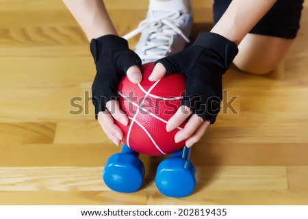 Horizontal image of female hands wearing workout gloves while lifting red weight ball of off blue dumbbells on wooden gym floor